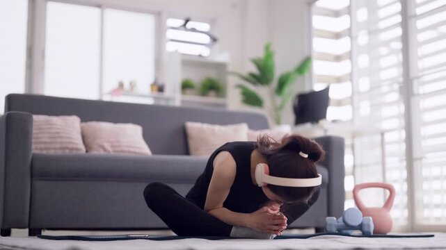 Asian adult woman perform home workout stretching exercise on yoga mat with headphones in modern living room promoting healthy lifestyle wellness flexibility