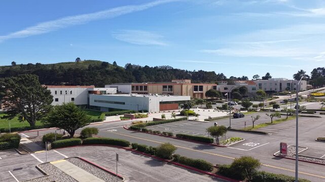 Rising aerial drone view flying towards the Skyline College campus buildings and empty parking lot in San Bruno, California, on a sunny day with blue sky and some clouds in the background