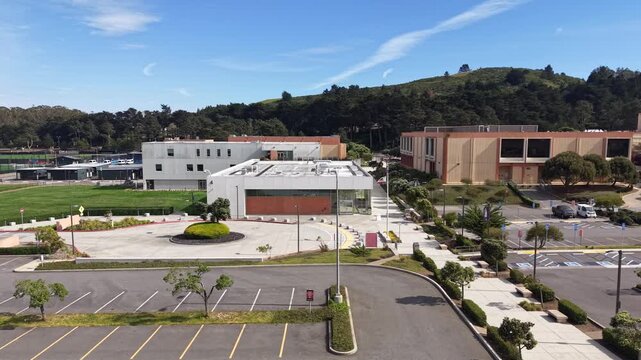 Sunny afternoon aerial perspective of Skyline College campus, featuring modern buildings, an empty parking lot, and green hillsides under a clear blue sky in San Bruno, California. Panning shot