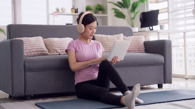 Asian woman using laptop computer wearing headphones enjoying leisure time study remote work activity home living room sofa floor
