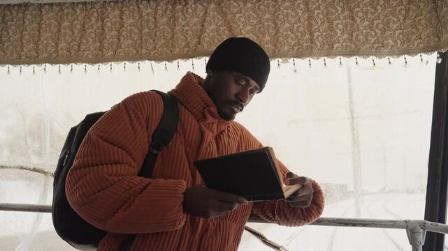 Black man reading and checking watch near transit shelter, backpack on shoulders, leafless trees beyond wet window, focused expression, turning pages, glancing at wristwatch, cold breath visible,