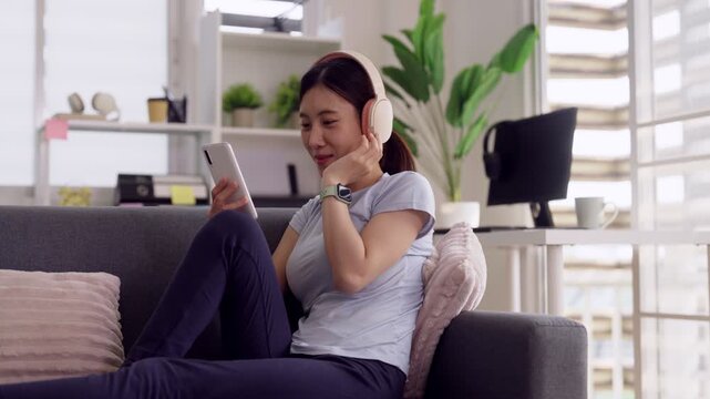 Asian adult woman enjoying music browsing smartphone relaxing couch home living room leisure entertainment content streaming technology modern lifestyle comfort