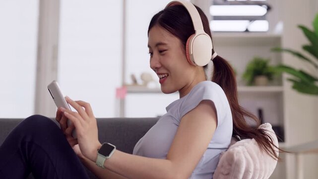Asian young adult woman wearing headphones uses smartphone enjoying music entertainment content home living room relaxed comfortable atmosphere