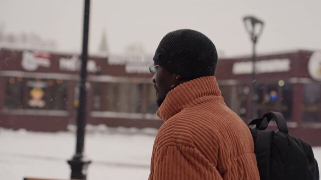 Black man in orange coat stands on snowy urban street, wearing beanie and backpack, glancing toward distant cafe and lamppost while light flakes fall frames suggest commuter hesitating, graduate