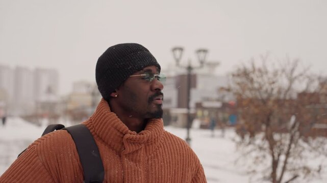 Black man orange sweater gazing across snowy plaza, independent traveler scouting route, backpack strap visible, lamp posts and pedestrians blurred, light snow falling, wide urban vista, hopeful