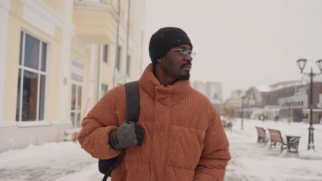 Black man walking snowy downtown street, wearing orange chunky coat, black beanie and glasses, carrying backpack, passing pastel storefronts and benches, snow flurries falling, lamppost silhouettes,