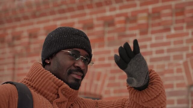 Black man in orange sweater waving with raised hand to block glare, smiling against brick wall candid urban portrait shows beanie, glasses, gloves, backpack and approachable commute vibe