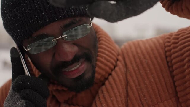 Black man outdoors in snow holding phone, wearing orange knit sweater, black beanie and mirrored glasses gloved hand shields face from falling flakes candid urban closeup capturing tense expression,