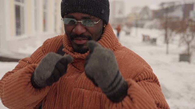 Black man shadowboxing on snowy sidewalk orange sweater knit beanie gloves sunglasses backpack dynamic jabs hooks playful training falling snow gritty urban energy candid exercise session