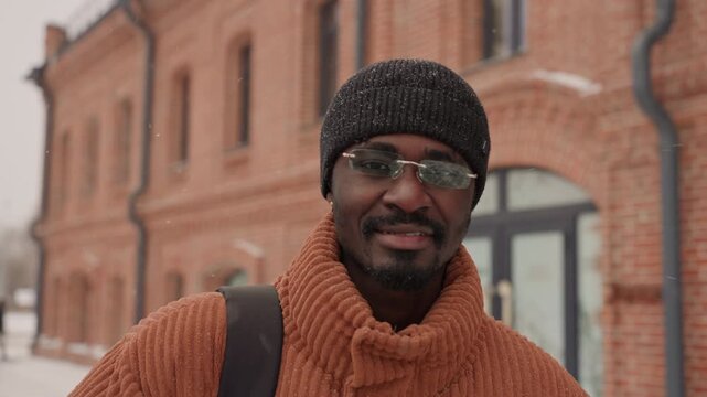 Smiling man near brick building in winter city, warm rust sweater and knit hat with snowflakes, backpack strap visible, candid gaze to camera by doorway and pedestrians, roles include tourist, local