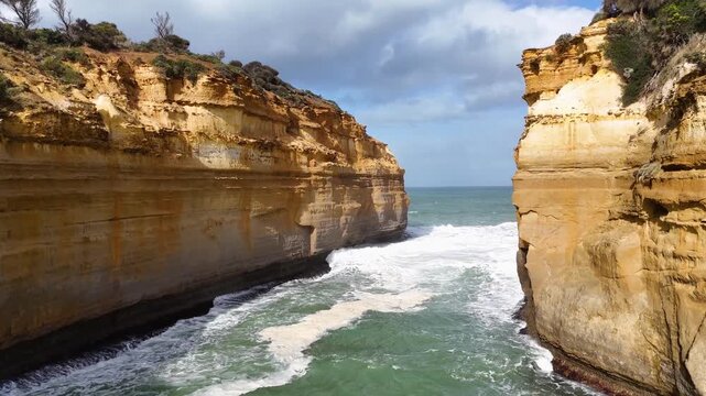 Cinematic aerial view of Loch Ard Gorge with turquoise ocean waves crashing against limestone cliffs in Australia.