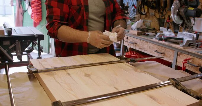 Carpentry workshop. Furniture assembly. Male joiner cleaning wet excess glue with cloth from wood panel tightened with bar clamps.