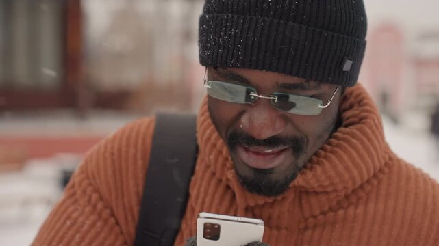 Black man walking in snowy city, knit sweater and beanie, talking on phone with thoughtful expression, backpack slung over shoulder, snowflakes falling, muted cold atmosphere, candid commuter portrait