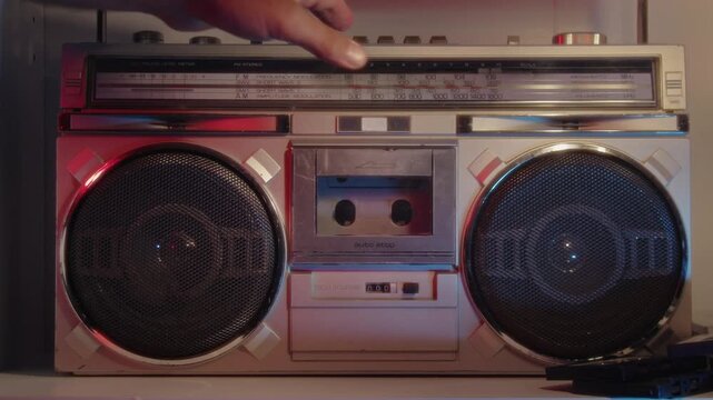 Macro close-up in neon-lit studio: hand loads audio cassette into vintage boombox, then presses play button. Warm analog sound begins, reels turn smoothly, strong 1980s retro atmosphere 4K.