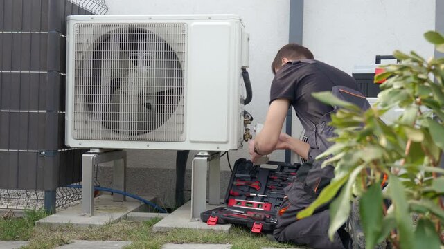 Technician performing maintenance on outdoor heat pump unit near residential building exterior. Man installing air source heat pump using tools. Concept of HVAC service and heating system repair