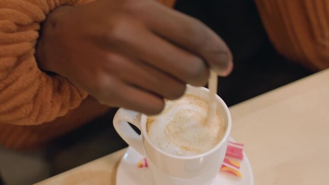Black hands stirring coffee with spoon, white mug on saucer and sugar packet, knit brown sleeve against wooden table, cinnamon dusting and gentle ripple motion, mindful pause during study session,