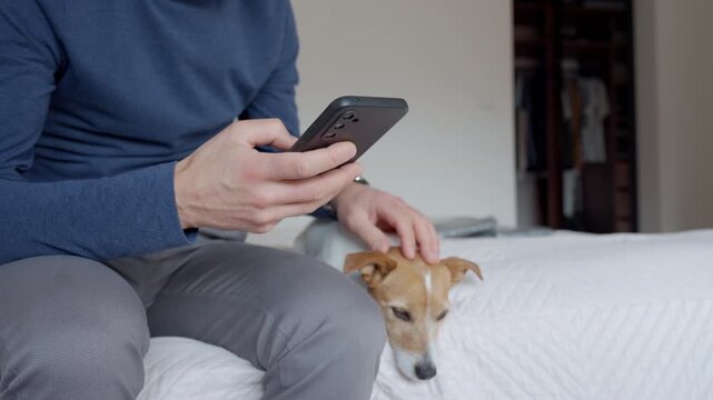 Man using smartphone while sitting on bed with small dog in home bedroom. Concept of mobile communication pet companionship home lifestyle and everyday technology