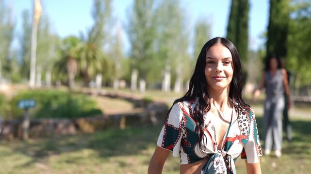 Young woman smiles, facing camera in park. She walks through sunlit grass, dressed in patterned top. Background figures play, slightly blurred, enjoying daylight. Natural setting features trees