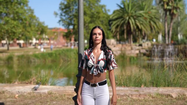 Woman leans against a lamppost in a park. She smiles softly at the camera. Her gaze shifts thoughtfully toward the horizon. She wears a bright, patterned top with white trousers