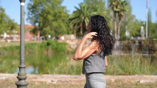 Woman stands by pond, back to camera. She runs hand through long, dark curls. Sunlight glints off greenery and water. She appears relaxed, enjoying outdoor moment. Ideal for wellness, travel