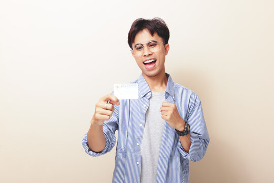 Excited young Indonesian man celebrating while holding ID card or KTP on isolated background.