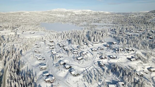 Wide aerial view of winter cabin retreat, Danebu, Valdres, Norway, frozen lake Damtjern, and alpine mountains in bright sun.