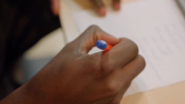 Black hands writing exam notes on paper with orangeblue pen, tense study atmosphere, closeup of handwriting and margins, student preparing under time pressure in library or classroom setting