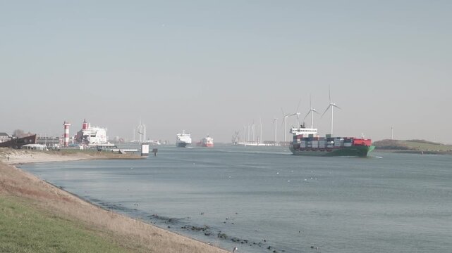 Large container ship sailing out of Rotterdam harbor near Hoek van Holland with calm water and wind turbines along the coastline in daylight