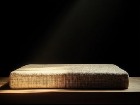 woven rectangular cushion on wooden surface bathed in warm spotlight with deep shadows, serene minimalist stillness and calm contemplative mood