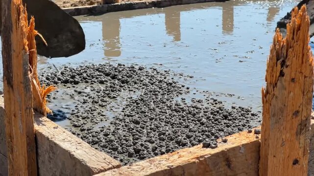 Construction worker using shovels to spread wet concrete mixture in wooden formwork on sunny day