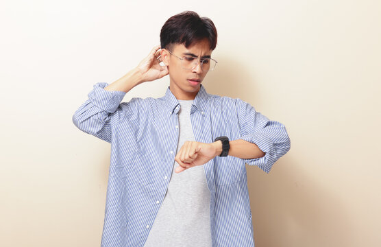 Confused young Asian man looking at wristwatch while scratching head with stressed gesture on isolated background. Time management and deadline concept.
