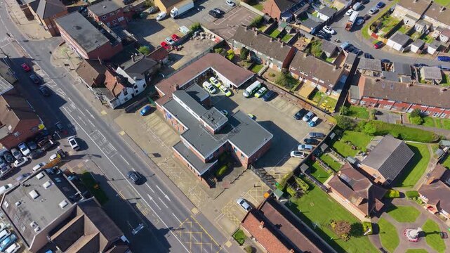 Cheshunt police station aerial descent establishing shot, revealing station building, marked vehicles, nearby streets and housing in Hertfordshire, England on a bright day.