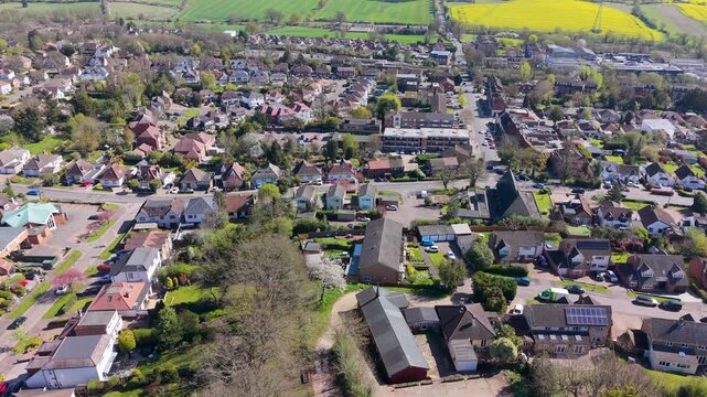 Cuffley, Hertfordshire aerial pan tracking parallel to Station Road, showing suburban homes, trees, gardens, fields, and village center on a bright spring day in England.