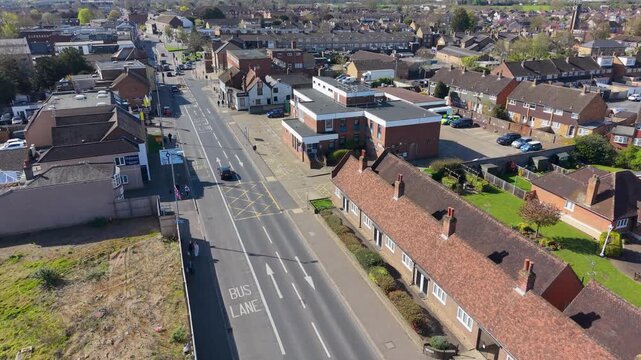 Cheshunt police station drone ascent establishing shot over Hertfordshire suburb with main road, almshouses, bus lane marking, rooftops, traffic and neighborhood buildings in daylight.