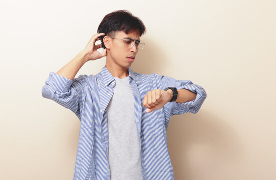 Confused young Asian man looking at wristwatch while scratching head with stressed gesture on isolated background. Time management and deadline concept.