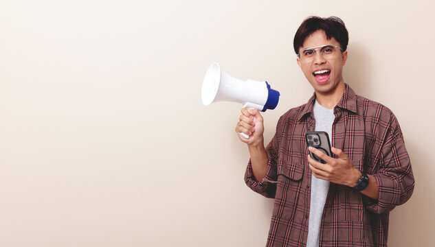 Excited young Asian man holding smartphone and shouting into a megaphone for digital promotion on isolated background.