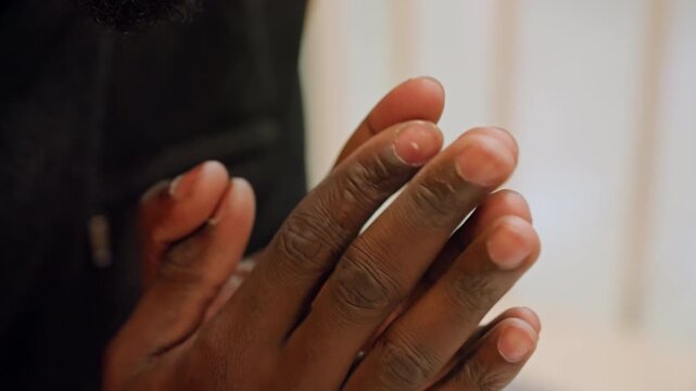 Closeup black man hands praying intimate indoor sequence showing hands clasp, gentle rubbing between fingers, textured skin, short nails, dark jacket sleeve, soft natural light, contemplative grateful