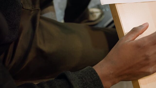 Black man touching parcel on table, seated at cafe counter, textured cardboard edge, relaxed forearm and wrist, casual clothing, anticipatory gesture, soft interior lighting, candid close detail