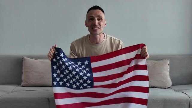 An enthusiastic American sports fan holding the United States national flag screams with joy and celebrates a goal while watching a football match at home