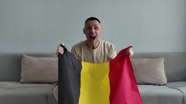 An enthusiastic Belgian sports fan holding the Belgian national flag shouts with joy and celebrates a goal while watching a football match at home