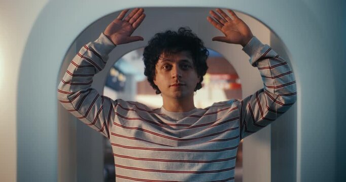 Young Man With Curly Hair in Striped Sweatshirt Stands Inside Modern Millimeter Wave Body Scanner. Guy Holds Hands Up as Instructed by Security Protocols. Standard Procedure of Aviation Security.