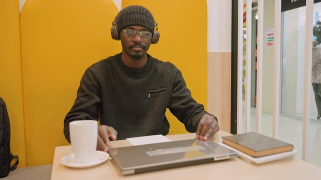 Man closing laptop at cafe table, yellow booth backdrop, headphones and coffee mug, notebook and pen beside computer, focused freelancer organizing laptop into backpack, soft natural light and tidy