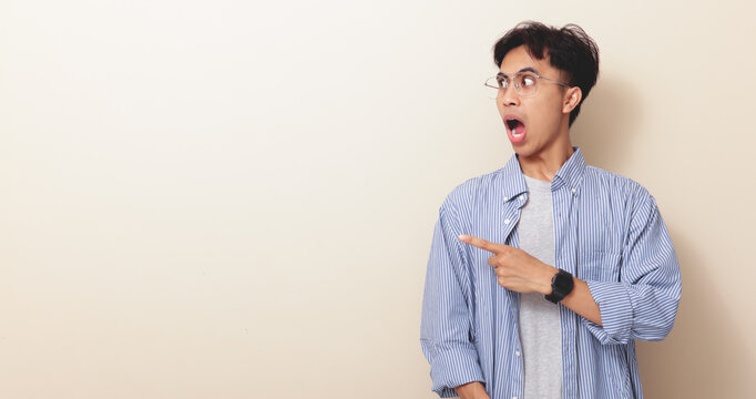 Funny young Asian man with surprised expression pointing sideways on isolated background.