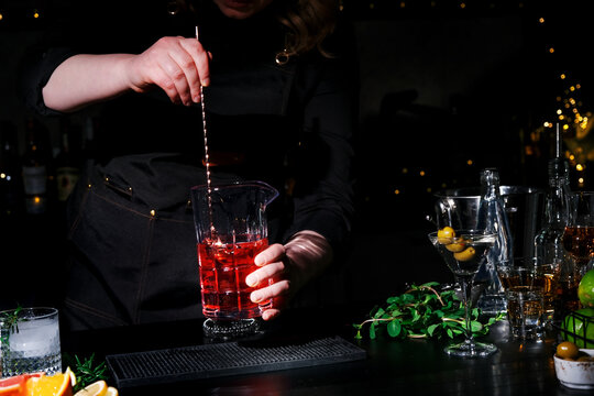 Woman bartender with bar spoon and red Manhattan cocktail in mixing glass. Professional mixology and drink dilution control.