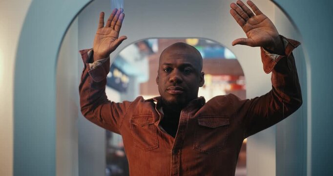Black Man in Terracotta Button Down Shirt Stands Inside State of Art Millimeter Wave Scanner. Male Calmly Raises Hands as Part of Mandatory Pre Flight Screening Process. International Travel Safety.