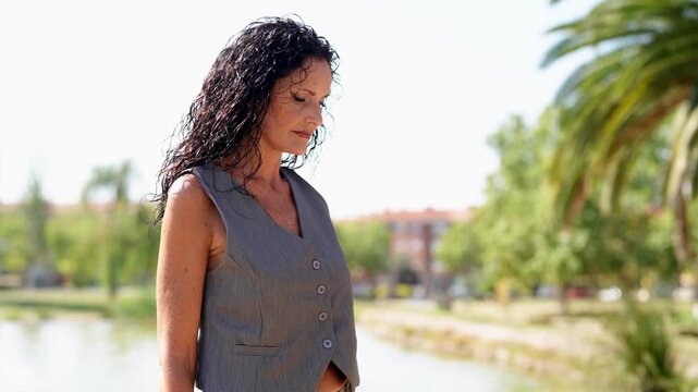 Woman stands by water, hand on head, calm expression. Sunlit park with trees and distant buildings. She shifts stance, arms adjusting, natural grace. Outdoor scene, relaxed, elegant, tranquil moment