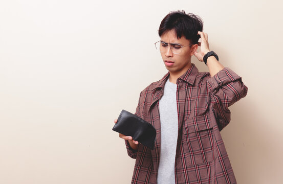 Confused young Asian man holding empty wallet with worried expression on isolated background.