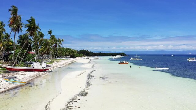 Scenic view of a white sand beach lined with palm trees and boats on clear blue water in Panglao, Philippines. Bright sunny day with calm sea and vibrant tropical atmosphere.