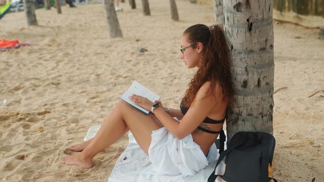 Caucasian woman journaling at sandy beach by palm trunk, focused creative retreat vibe, blue notebook and pen, white skirt, backpack at side, barefoot on towel, soft daylight, distant swimmers,