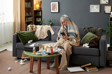 Wide shot of senior Caucasian woman checking smartphone while sitting wrapped in blanket among medicines and tissues, showing illness recovery. Useful for healthcare, flu season, home care marketing © pressmaster